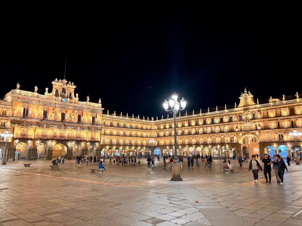 Vue nocturne de la Plaza Mayor de Salamanque, en Espagne, avec des bâtiments historiques baignant dans une lumière dorée. Si vous prévoyez de visiter Salamanque en 3 jours, cette place vibrante et animée la nuit est à voir absolument sous les lampadaires scintillants.