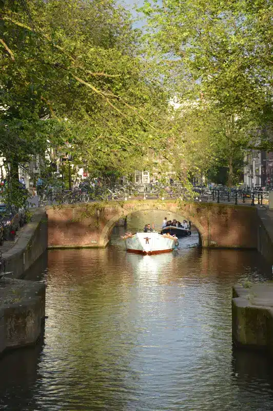 Un petit bateau avec des passagers passe sous un pont bas en briques, sur un canal bordé d'arbres à Amsterdam ; la lumière du soleil filtre à travers le feuillage et des vélos sont garés le long des berges : une scène typique pour ceux qui visitent Amsterdam.