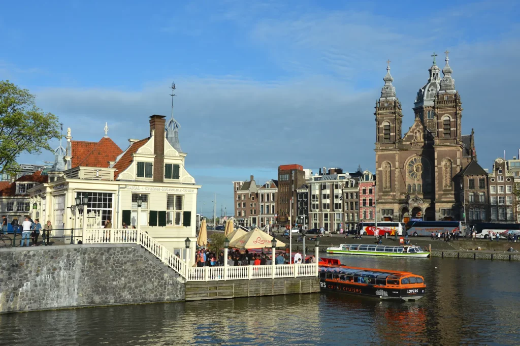 Un canal d'Amsterdam avec un bateau-mouche, des bâtiments historiques, une terrasse de café et les tours jumelles de la basilique Saint-Nicolas sous un ciel bleu : un incontournable pour tous ceux qui souhaitent visiter Amsterdam.