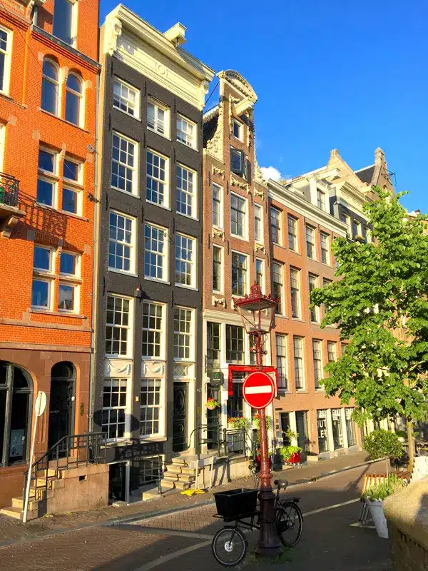 Une rangée de bâtiments colorés, hauts et étroits, le long d'une rue ensoleillée, avec un panneau « Sens interdit » rouge et blanc, un vélo garé près d'un lampadaire et des arbres verts au premier plan.