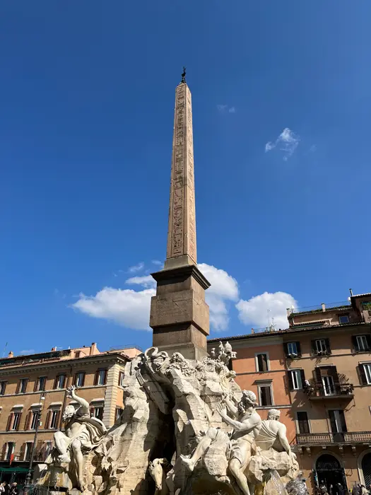 Un grand obélisque s'élève au-dessus d'une fontaine ornée de statues de personnages et de chevaux, sur fond de ciel bleu et entourée de bâtiments historiques.