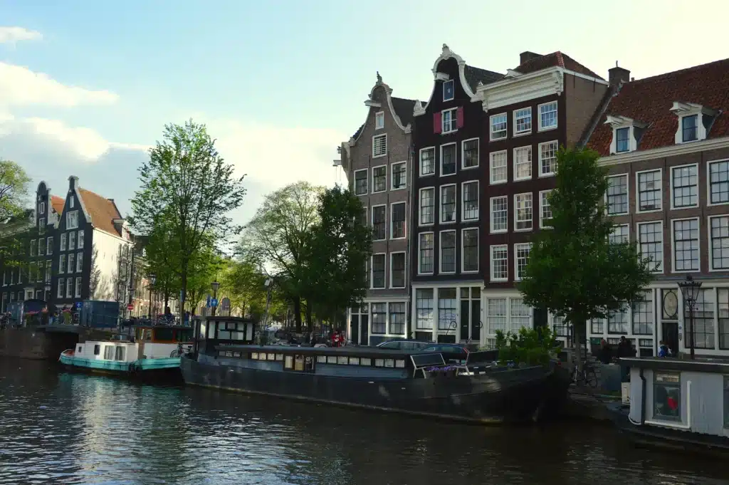 Canal à Amsterdam bordé de maisons hollandaises traditionnelles étroites, d'arbres et de bateaux amarrés au bord de l'eau, sous un ciel partiellement nuageux.