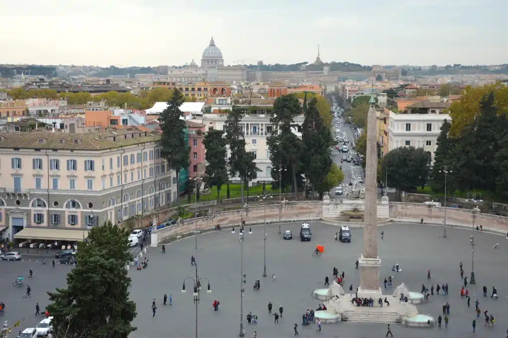 Vue de la Piazza del Popolo à Rome, avec un obélisque au centre, entouré de personnes et de bâtiments historiques, la basilique Saint-Pierre étant visible à l'arrière-plan.