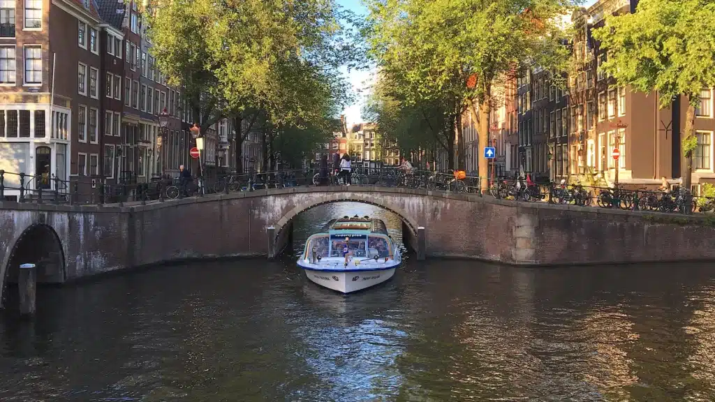 Un bateau-mouche blanc passe sous un pont en briques sur un canal bordé de grands arbres et de bâtiments historiques à Amsterdam ; on aperçoit des passants et des vélos sur le pont.