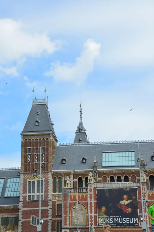 La photo montre la façade du Rijksmuseum à Amsterdam, avec ses tours en briques richement décorées et une grande banderole publicitaire pour le musée, le tout sur fond de ciel bleu parsemé de légers nuages.