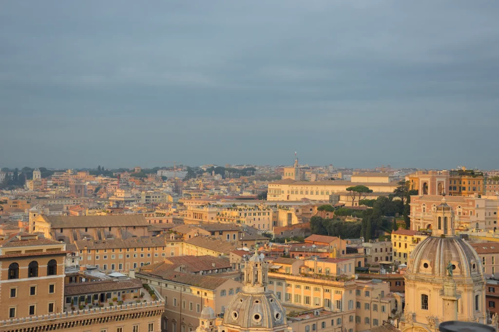 Vue panoramique de Rome au coucher du soleil, avec des bâtiments historiques, des églises à coupole et une douce lumière dorée illuminant la ville sous un ciel partiellement nuageux.