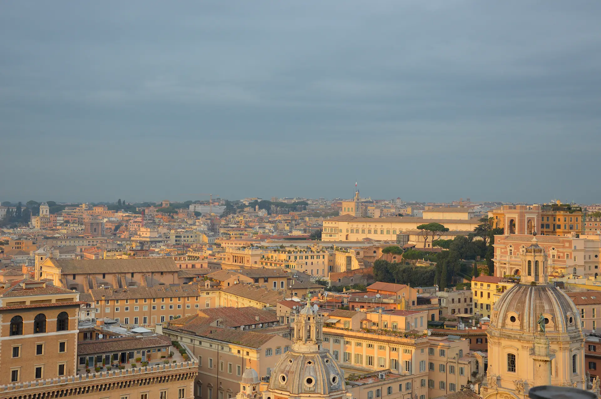 Vue panoramique de Rome au coucher du soleil, avec des bâtiments historiques, des églises à coupole et une douce lumière dorée illuminant la ville sous un ciel partiellement nuageux.