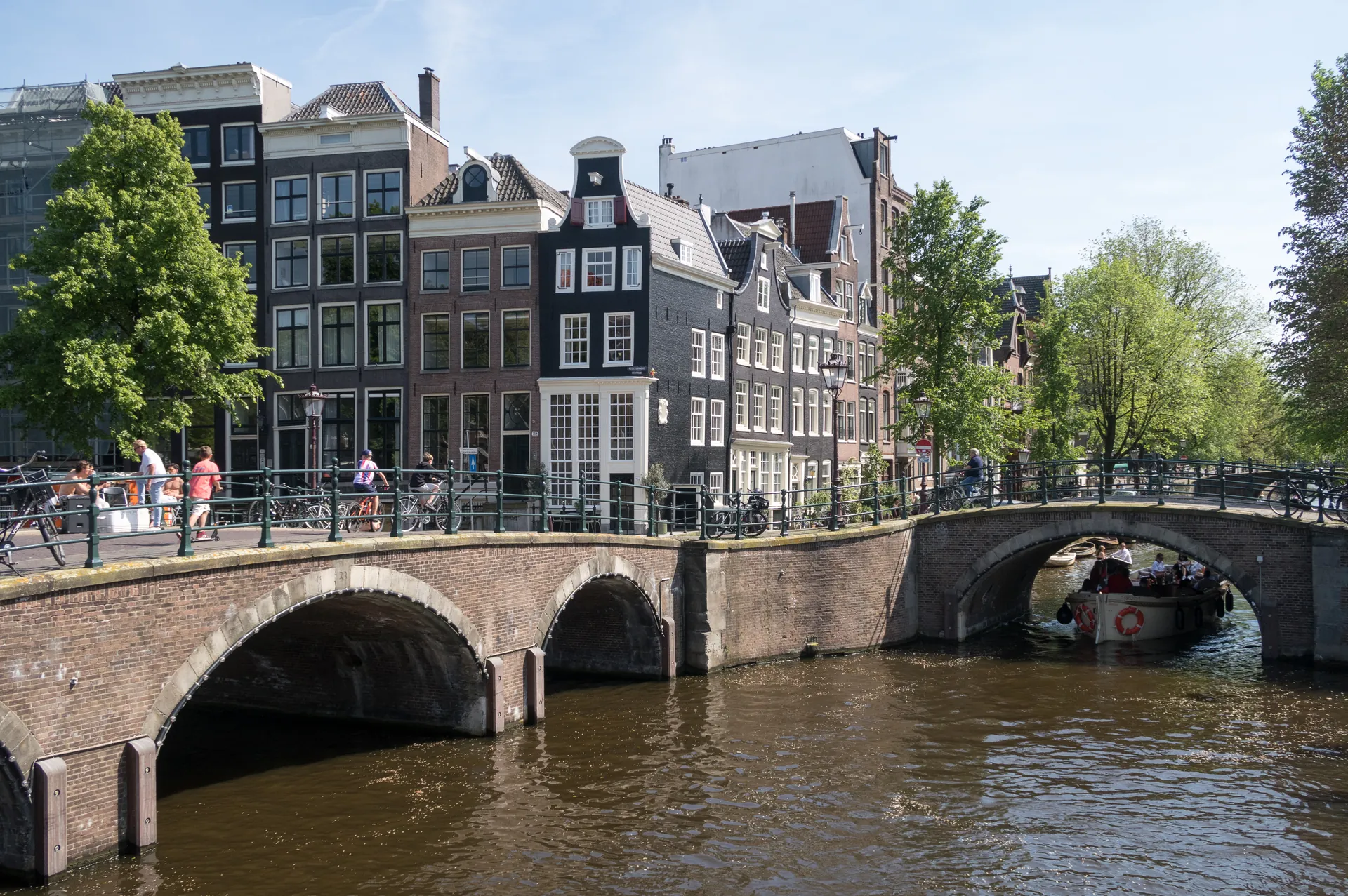 Un canal à Amsterdam, avec ses ponts en briques voûtés, ses maisons historiques étroites, ses arbres verdoyants, ses promeneurs et ses cyclistes, et un bateau passant sous le pont par une journée ensoleillée : l'image parfaite pour ceux qui se demandent « quand aller à Amsterdam ».