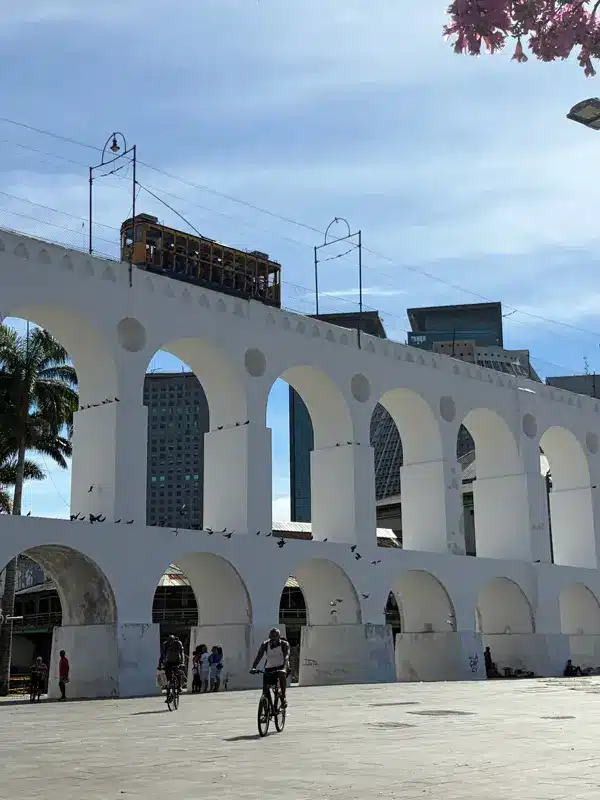 Des cyclistes passent sous les arches blanches de l'aqueduc de Lapa à Rio de Janeiro, avec un tramway jaune qui circule au-dessus et les immeubles de la ville en arrière-plan, sous un ciel bleu.
