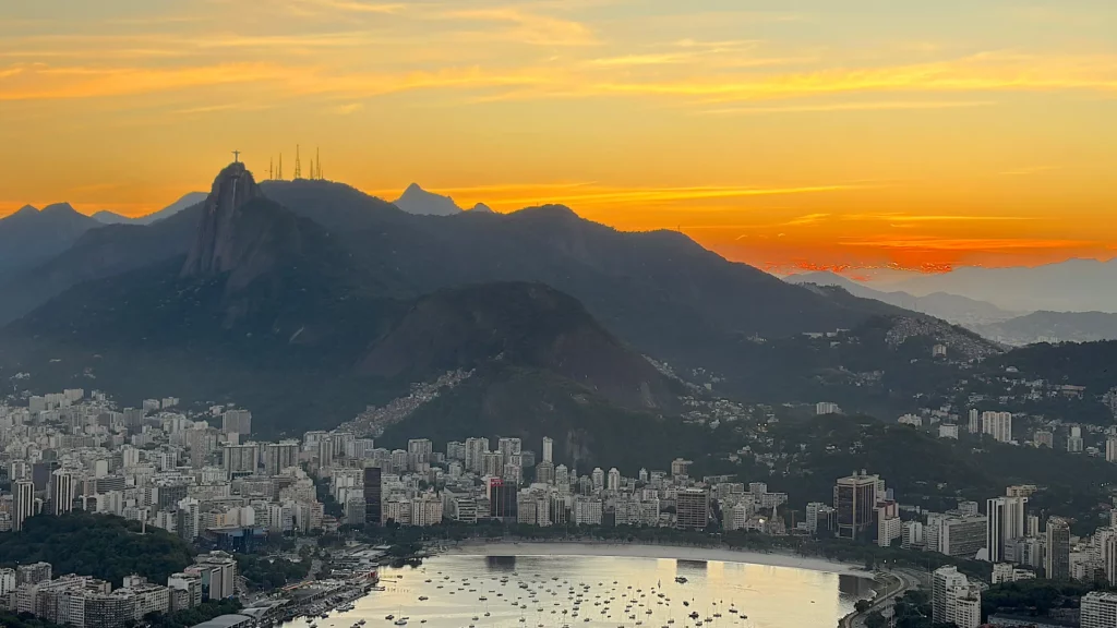 Un coucher de soleil éclatant sur Rio de Janeiro attend ceux qui visitent la ville, avec ses montagnes, ses immeubles et sa baie parsemée de bateaux ; le ciel se pare de teintes orange et jaunes au-dessus des sommets se découpant en silhouette.