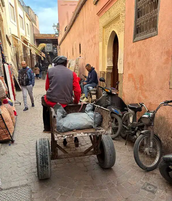 Une rue étroite où l'on aperçoit un homme poussant une charrette, des passants, des motos garées contre un mur aux teintes rosées, et des boutiques alignées de part et d'autre. La rue dégage une atmosphère historique, typique de la vieille ville — exactement le genre de scène pleine de charme que l'on peut découvrir en visitant la médina de Marrakech.