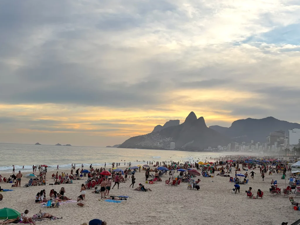 Une plage bondée au coucher du soleil, où les gens se détendent, avec des parasols éparpillés sur le sable, des vagues venant s'échouer sur le rivage, et en arrière-plan, des montagnes caractéristiques ainsi que les immeubles de la ville, le tout sous un ciel partiellement nuageux — une ambiance qui illustre parfaitement pourquoi tant de gens se demandent « quand aller à Rio de Janeiro » pour vivre une expérience inoubliable.