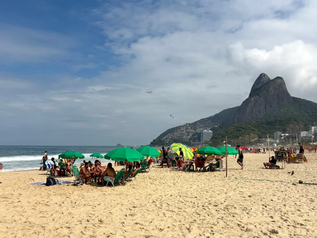 Des personnes se détendent sous des parasols verts sur la plage d'Ipanema, avec en toile de fond les vagues de l'océan et les montagnes au loin, sous un ciel partiellement nuageux, créant ainsi l'atmosphère sereine que recherchent souvent ceux qui se demandent « Où dormir à Rio de Janeiro ? ».