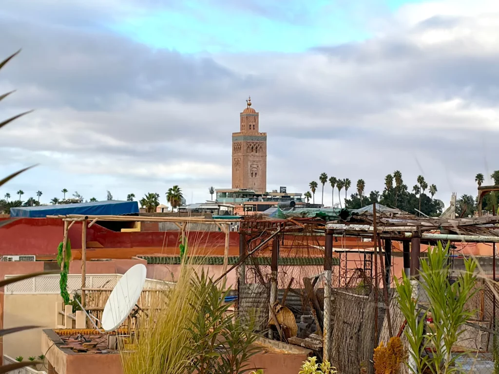 Une vue depuis un toit, avec des plantes et une antenne parabolique au premier plan, et la tour de la mosquée Koutoubia se dressant à l'arrière-plan sous un ciel nuageux à Marrakech, au Maroc : une vue idéale pour ceux qui souhaitent visiter la médina de Marrakech et s'imprégner de son atmosphère animée.
