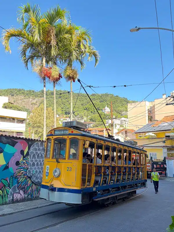 Un tramway jaune circule dans une rue avec des passagers à bord, longeant des œuvres d'art urbain colorées et de grands palmiers sous un ciel bleu limpide, dans un quartier urbain ensoleillé — une scène qui illustre parfaitement l'ambiance animée de la ville pour tous ceux qui se demandent « que faire à Rio de Janeiro en 3 jours ».