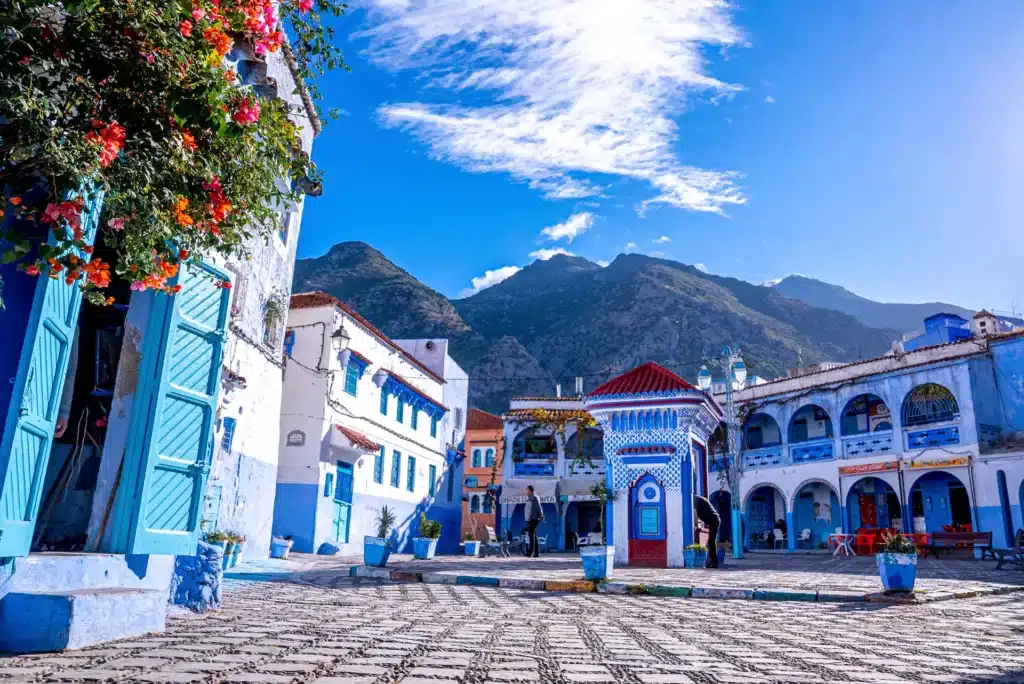 Une place de ville animée, avec ses bâtiments bleus et blancs, ses fleurs colorées, les montagnes en arrière-plan et un ciel d'un bleu éclatant parsemé de nuages, vous invite à découvrir Chefchaouen et à vous laisser séduire par son charme inoubliable.