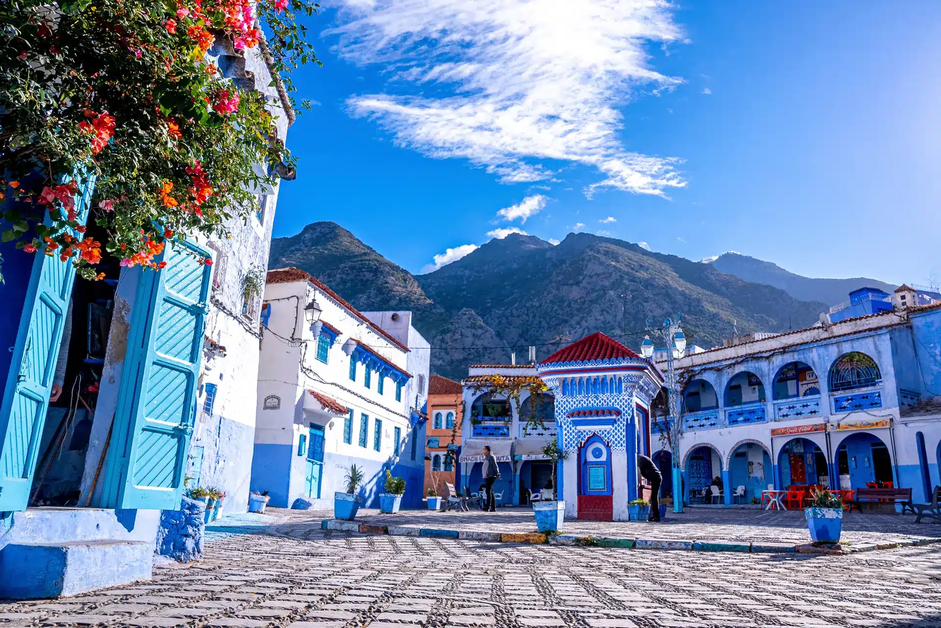 Une place de ville animée, avec ses bâtiments bleus et blancs, ses fleurs colorées, les montagnes en arrière-plan et un ciel d'un bleu éclatant parsemé de nuages, vous invite à découvrir Chefchaouen et à vous laisser séduire par son charme inoubliable.