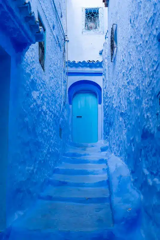 Ruelle étroite aux murs peints en bleu, avec des marches et une porte bleue, à Chefchaouen, au Maroc. Une petite fenêtre à barreaux surplombe la porte, et la scène est baignée d’une lumière naturelle éclatante — une invitation charmante à visiter Chefchaouen et à découvrir par soi-même son charme unique.