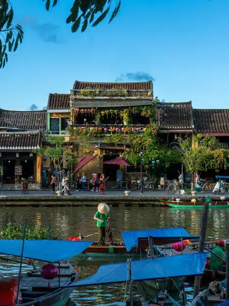 Une personne coiffée d'un chapeau conique rame dans une petite barque en bois sur une rivière, devant des bâtiments historiques : une scène typique pour ceux qui visitent Hoi An.