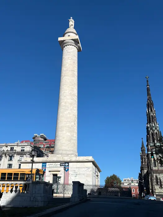 Le Washington Monument de Baltimore, une haute colonne de pierre surmontée d'une statue, se dresse à côté d'une église de style gothique et d'un bus scolaire jaune : un site à ne pas manquer si vous visitez Baltimore en trois jours.