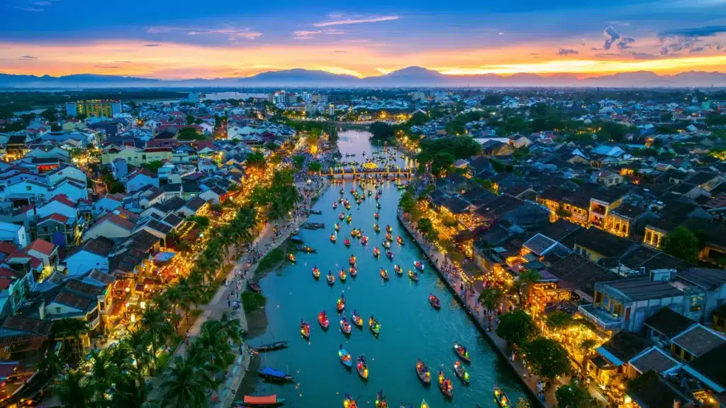 Vue aérienne au coucher du soleil sur les bateaux naviguant sur la rivière à Hoi An, au Vietnam, au milieu des bâtiments illuminés et des berges bondées : un spectacle magnifique pour ceux qui visitent Hoi An.